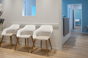 Wartebereich der Schmerztherapiepraxis Dr. Mesa in Iserlohn-Letmathe, mit weißen Stühlen, cremefarbenen Sitzkissen und Holzfüßen, Blick Richtung Anmeldebereich und Behandlungsräume mit türkisblauen Wänden – gestaltet von Grothe Büro & Objekteinrichtung, Fotografiert von Susanne Beimann.