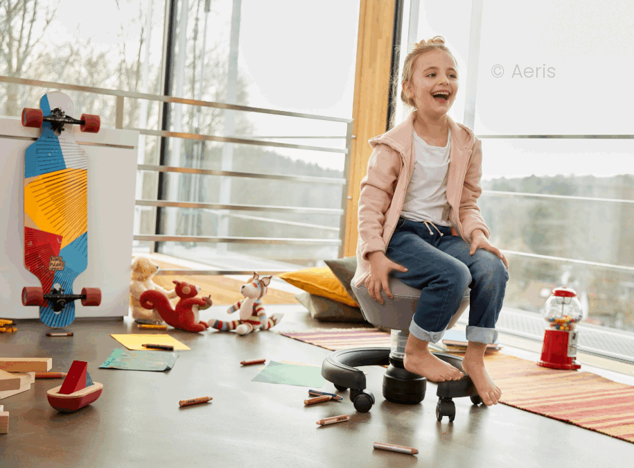 Ein Mädchen im Grundschulalter sitzt lachend auf einem Aeris Swoppster, einem ergonomischen Schreibtischstuhl für Kinder. Sie befindet sich in einem Kinderzimmer mit großen Fensterfronten.