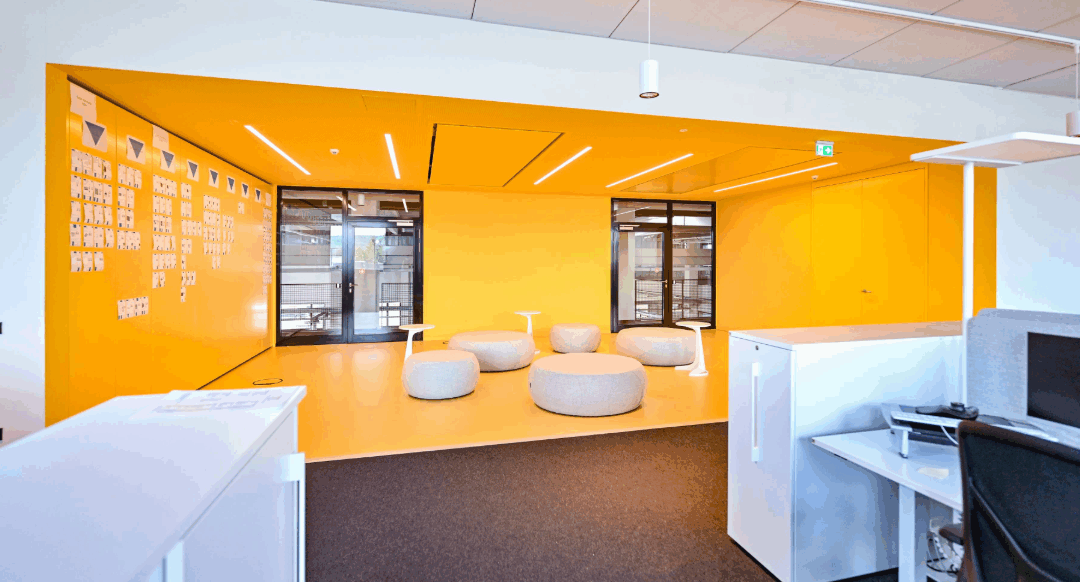 Ansicht eines Meeting Points in der Firma Bürkert, der als offener Bereich mit orangefarbenenen Wänden, Decke und Boden gestaltet wurde. Im Raum sind flexible Sitzmöbel für flexibles Arbeiten im Team