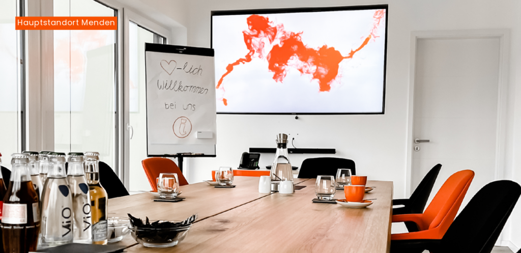Heller Konferenzraum am Hauptstandort Menden: Großer Eichenholz-Tisch ausgestattet mit modernen Konferenzstühlen in Schwarz und Orange, ergänzt durch professionelle Präsentationstechnik für kreative Teamarbeit.